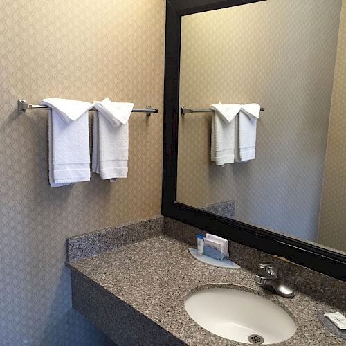 The image shows a bathroom with a granite countertop, a sink, a large mirror, two white towels on a rack, and toiletries on the counter.