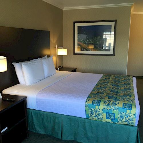 A neatly made bed in a well-lit hotel room with a side table, lamp, wall art, and clean, simple decor, ending the sentence.