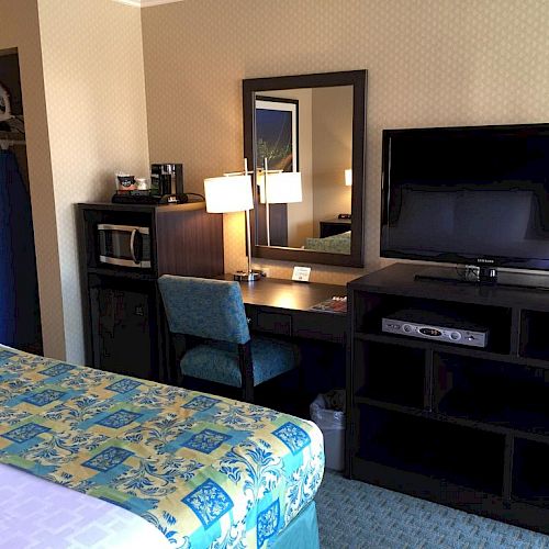 A hotel room includes a bed, TV, desk with a mirror, chair, microwave, and wardrobe. The overall decor is modern and simple.