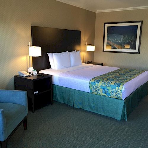 A hotel room with a neatly made bed, two bedside tables with lamps, a blue armchair, and a framed picture on the wall.