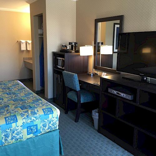 A hotel room with a bed, a desk with a chair, a mirror, a TV on a stand with storage shelves, and a bathroom area with towels hanging.