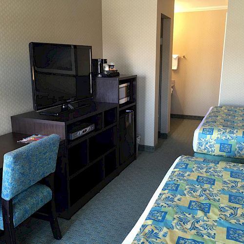 A hotel room with two beds, a TV on a stand, a desk with a chair, and a partially visible bathroom entrance in the background.