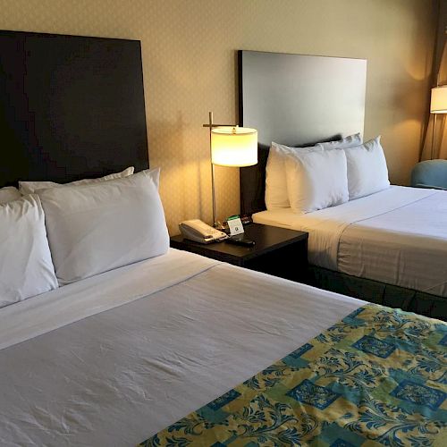 The image shows a hotel room with two neatly made beds, a bedside table with a lamp and phone, and a window with light coming through the curtains.