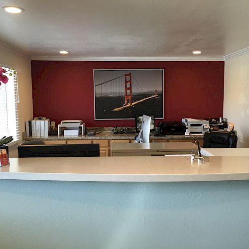 A reception area with a white counter, office equipment, a clock, pink flowers, and a picture of a bridge on a red wall in the background.