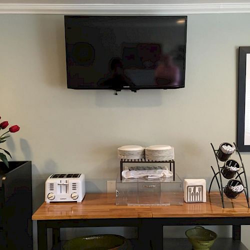 A wall-mounted TV above a small table with bowls, a towel rack, toaster, and a stack of napkins. Two framed pictures and a vase with red flowers.