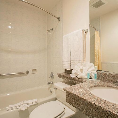 A clean bathroom with a bathtub, shower curtain, sink, towel rack, mirror, and toilet. The countertop has towels and toiletries.