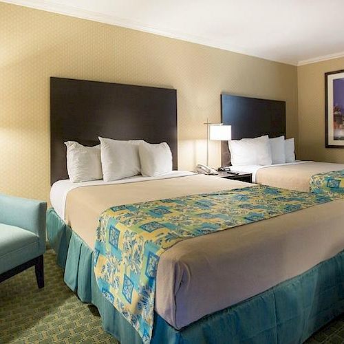 A hotel room featuring two beds with teal and yellow bedding, a light blue armchair, a modern lamp, and a framed picture on the wall.