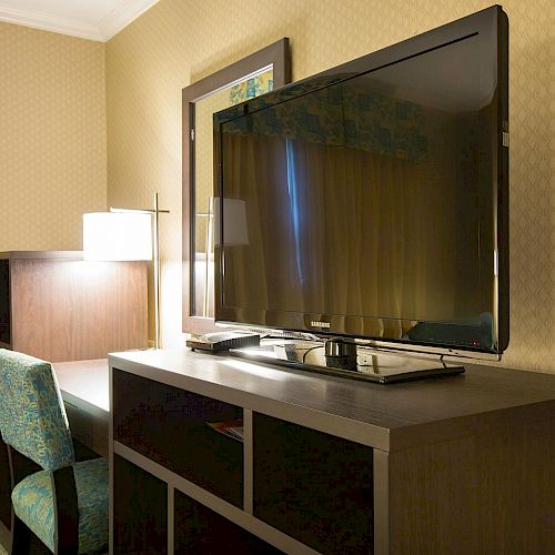 The image shows a hotel room with a large flat-screen TV on a wooden cabinet, a desk with a lamp, and a chair with a patterned fabric.