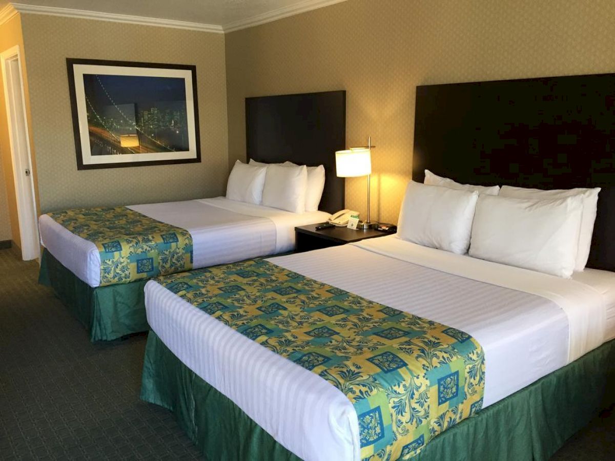 The image shows a hotel room with two neatly made double beds, a bedside table with a lamp and phone, and a framed picture on the wall.