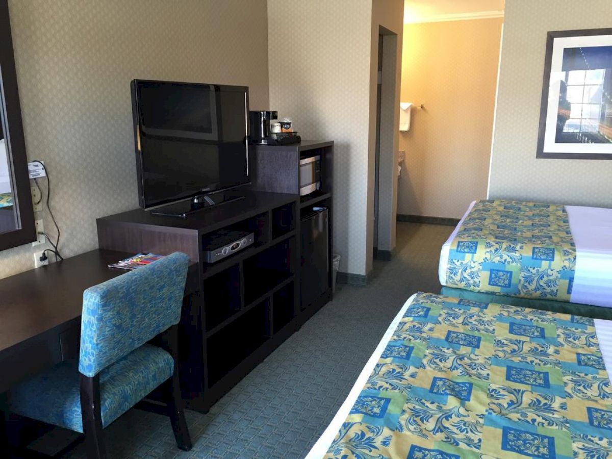 The image shows a hotel room with two beds, a desk and chair, a flat-screen TV, and an open doorway to a bathroom.