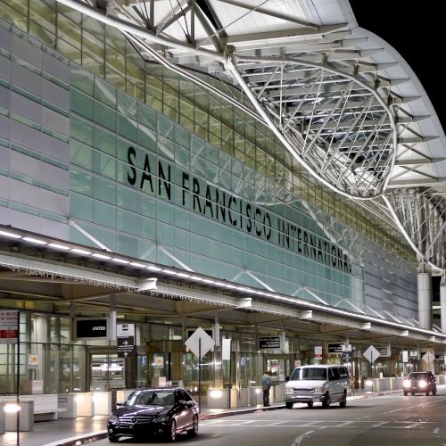 The image shows the exterior of San Francisco International Airport at night, featuring its illuminated signage and several vehicles in front.