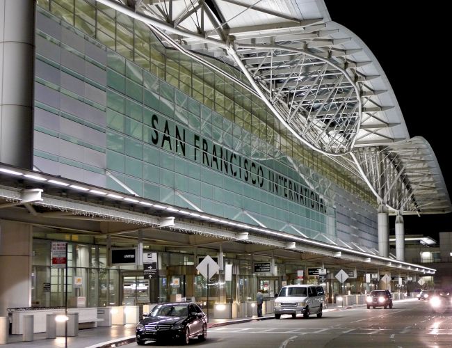 The image shows the exterior of San Francisco International Airport at night, featuring its illuminated signage and several vehicles in front.