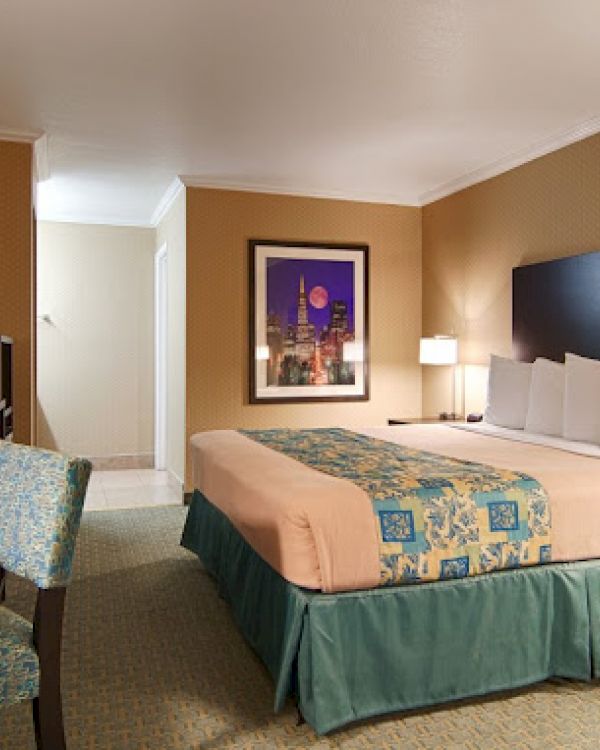 A hotel room with a large bed, nightstands, lamps, a desk with chair, a television, and a framed picture on the wall.