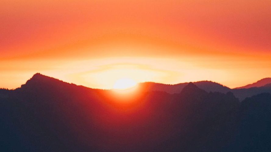 A vibrant sunset over a mountain range, with the sun setting behind dark peaks, casting a warm orange and red glow across the sky.