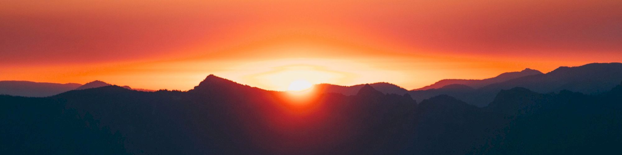 A vibrant sunset over a mountain range, with the sun setting behind dark peaks, casting a warm orange and red glow across the sky.