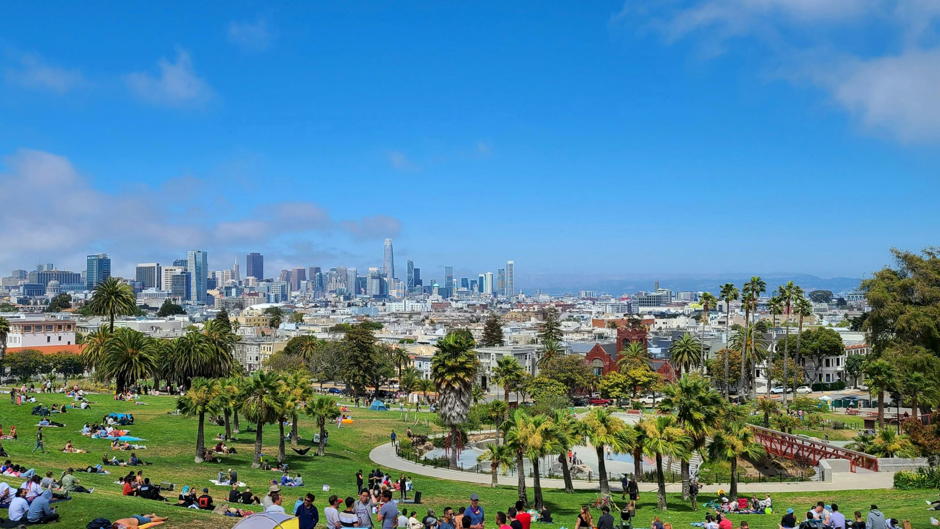 A park filled with people relaxing and enjoying the day, with a city skyline in the background under a partly cloudy sky.
