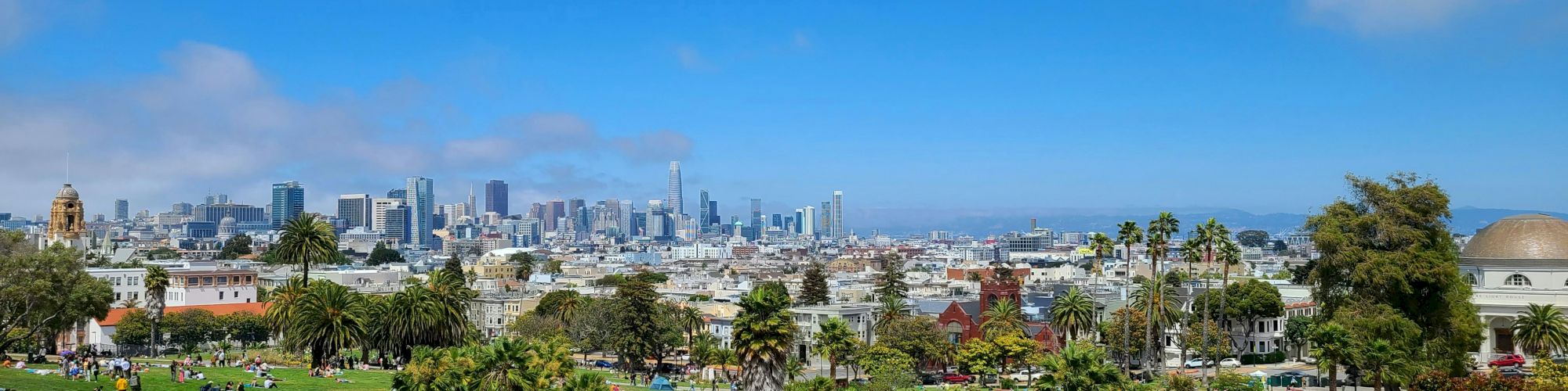 A park filled with people relaxing and enjoying the day, with a city skyline in the background under a partly cloudy sky.