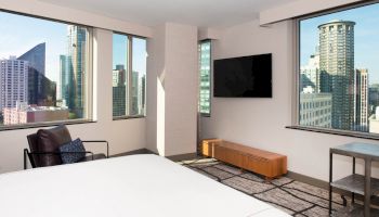 A modern hotel room with a city view, featuring large windows, a bed, a chair, a TV mounted on the wall, and a wooden bench.