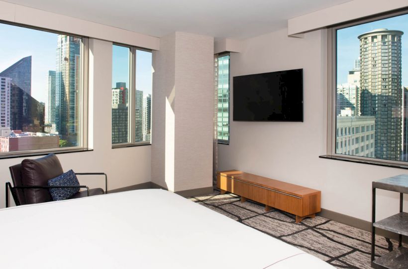 A modern hotel room with a city view, featuring large windows, a bed, a chair, a TV mounted on the wall, and a wooden bench.