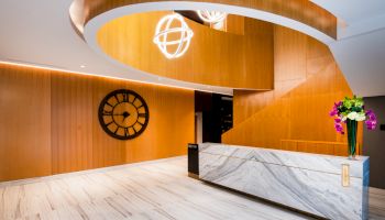 A modern lobby with a marble reception desk, wooden curved staircase, large clock on a wooden wall, and floral arrangement on the desk.