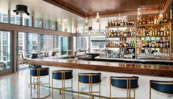 The image shows a stylish, modern bar with a wooden ceiling, a well-stocked bar shelf, blue barstools, and large windows offering city views.