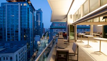 A modern urban balcony with seating overlooks a city street below. The interior appears to be a stylish office or restaurant with tables and chairs.