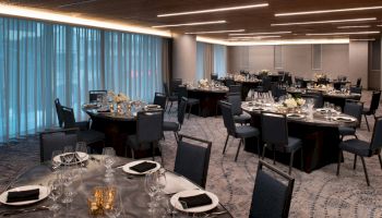 A banquet hall is set with round tables, black chairs, and elegant table settings, ready for an event, with dim overhead lighting.