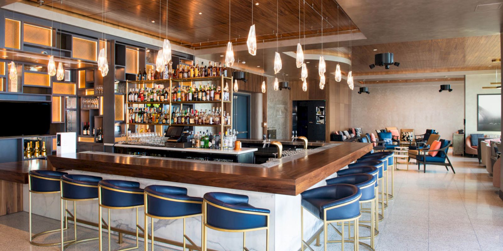 A modern bar with a wooden counter, blue velvet stools, hanging lights, and a well-stocked display of various liquor bottles behind the bar.