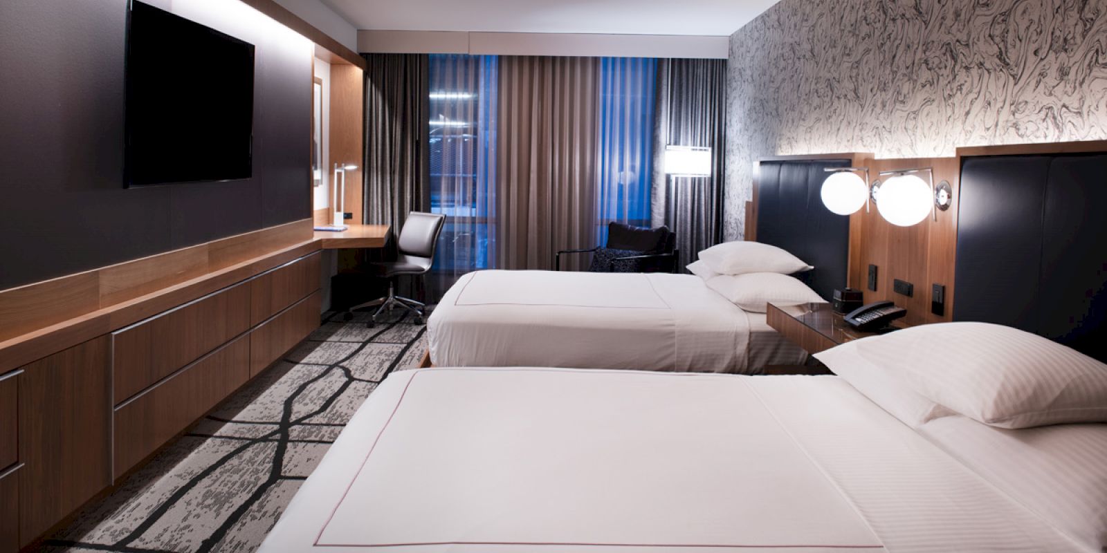 The image shows a modern hotel room with two neatly made beds, a wall-mounted TV, a desk with a chair, and bedside lamps.