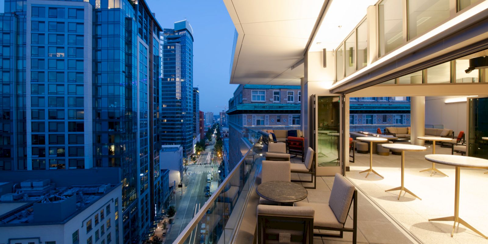 A modern rooftop terrace at dusk with outdoor seating, overlooking a cityscape with tall buildings and a street below, under a clear evening sky.