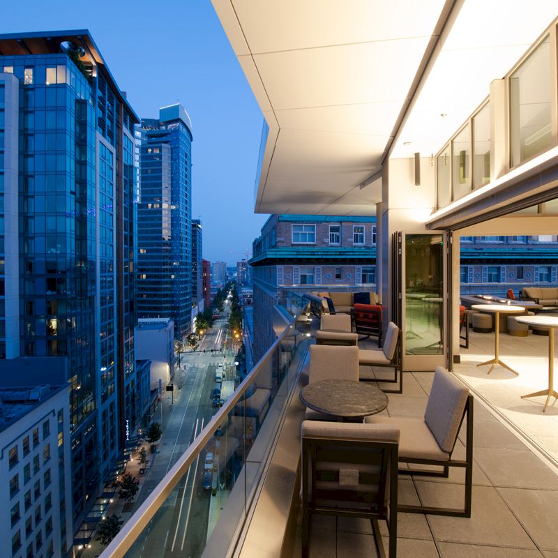 A modern cityscape featuring high-rise buildings at dusk, viewed from a stylish balcony with tables and chairs on the street below.