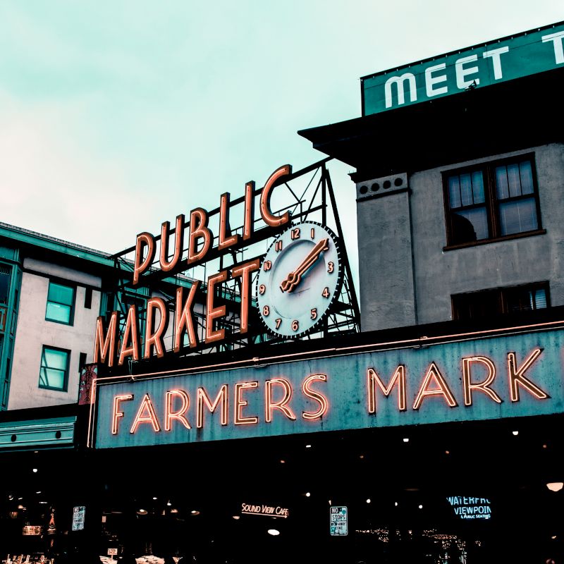 The image shows a neon sign for a Public Market and Farmers Market, with a vintage clock in a bustling area with nearby buildings.