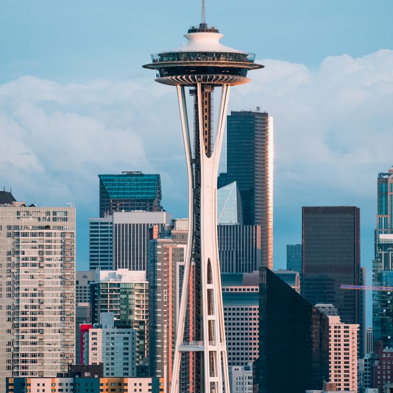 The image shows the Space Needle surrounded by various skyscrapers and buildings, set against a backdrop of blue sky and white clouds.
