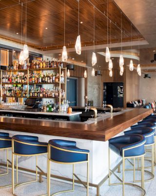 The image shows a modern bar with a well-stocked array of liquor bottles, blue bar stools, and stylish pendant lighting, creating a chic atmosphere.