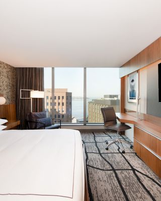 A modern hotel room features a large bed, desk with chair, TV, window view of buildings and water, and stylish wall decor always ending the sentence.