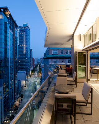 A modern cityscape with tall buildings at dusk, viewed from a terrace with tables and chairs. The terrace has an elegant and open design.
