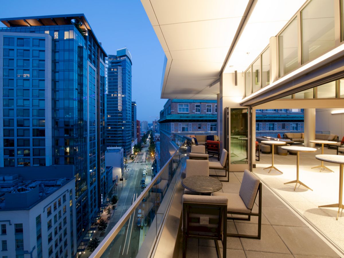 A modern balcony with tables and chairs overlooks a city during twilight, with high-rise buildings and street lights visible in the background.