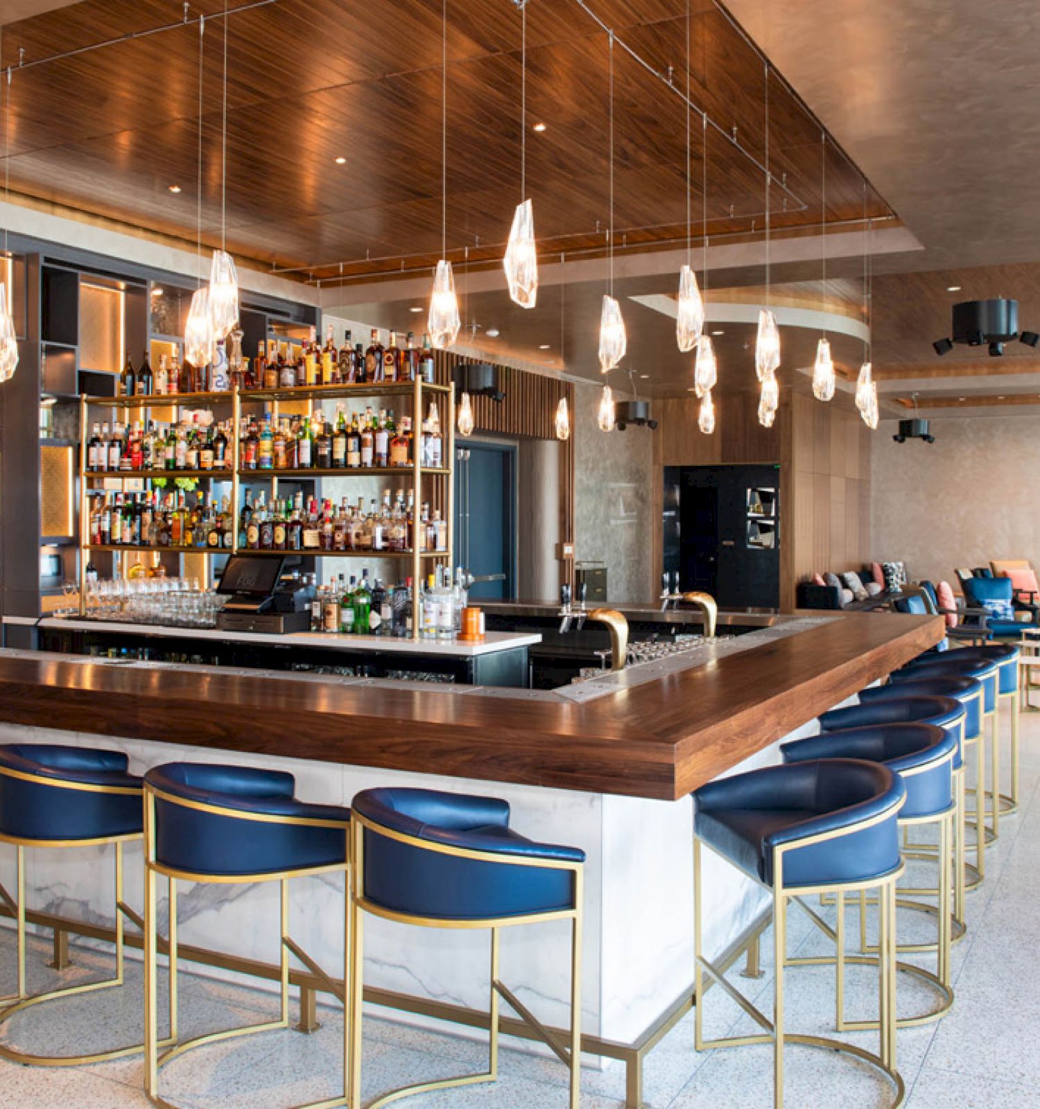 A modern bar area with a wooden counter, blue bar stools, ceiling lights, and shelves stocked with bottles, surrounded by seating areas.