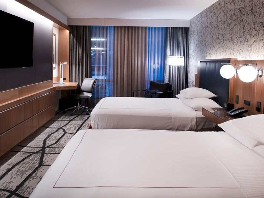 A modern hotel room features two beds, a wall-mounted TV, a desk with a chair, and stylish lighting, with large windows and patterned curtains.