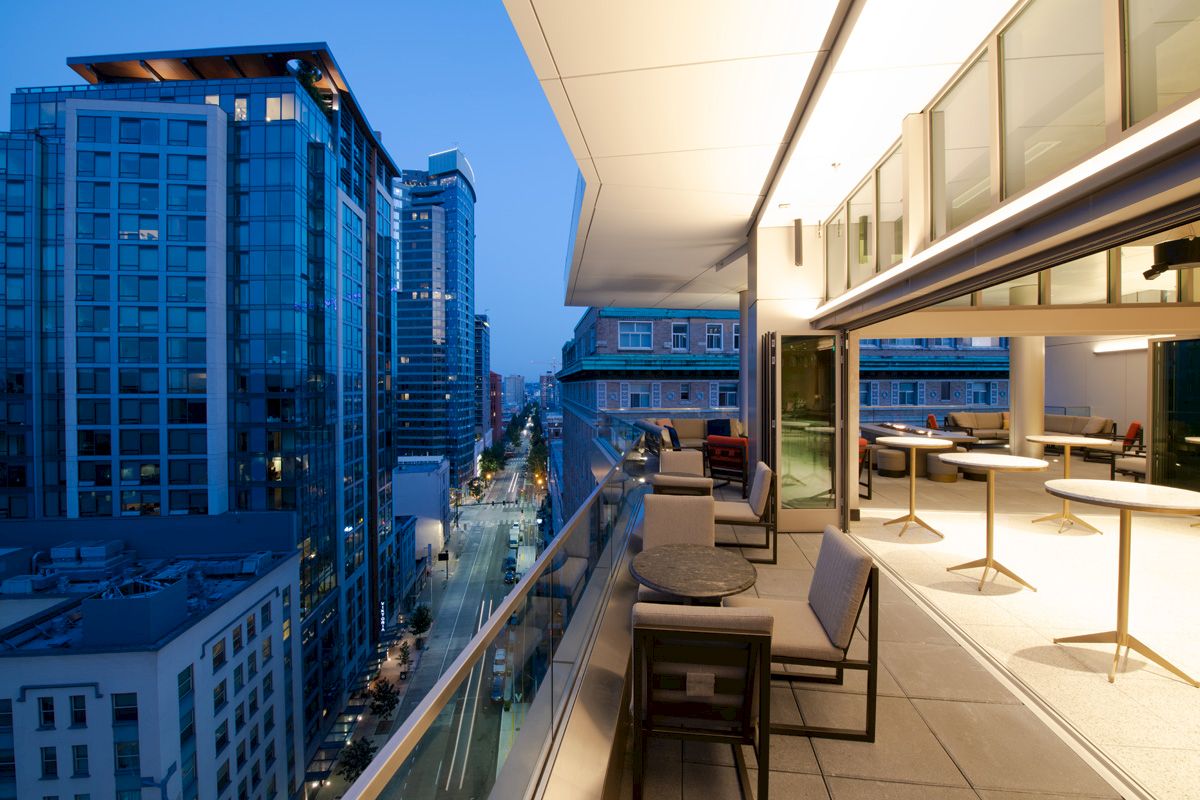 An urban balcony with tables, chairs, and city view at twilight. Adjacent buildings feature glass facades; streets below are illuminated.