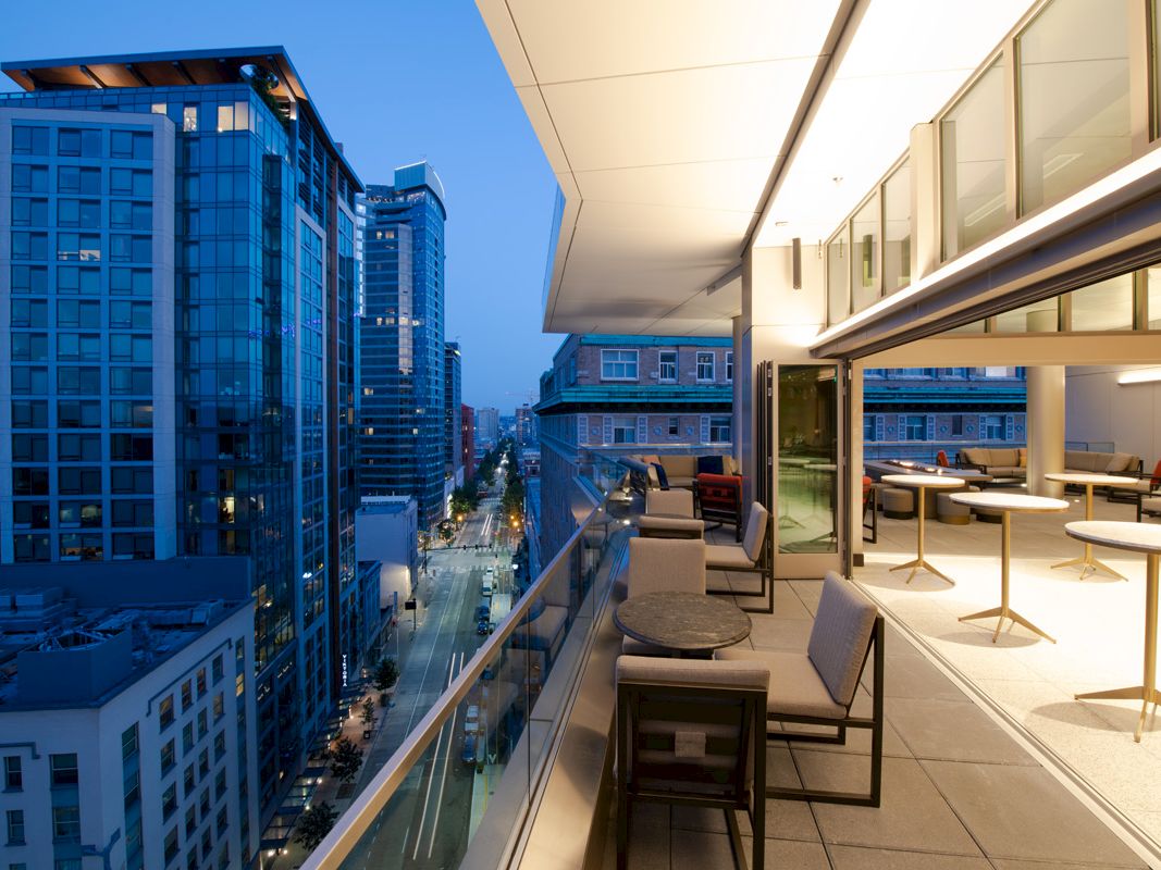 An urban balcony with tables, chairs, and city view at twilight. Adjacent buildings feature glass facades; streets below are illuminated.