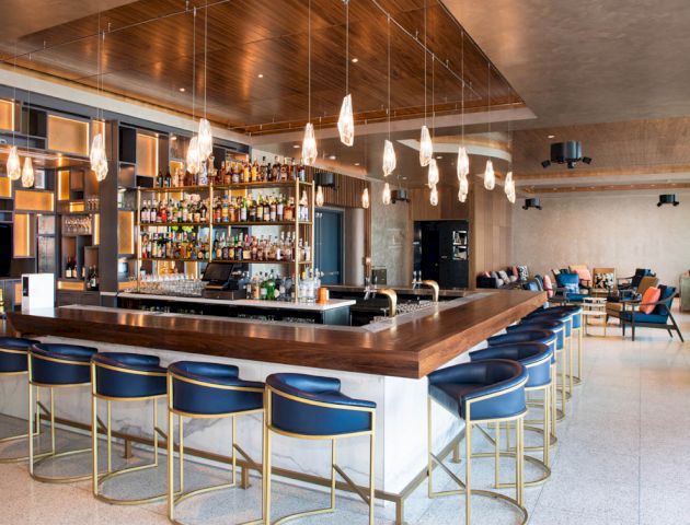 The image shows a modern bar with a U-shaped counter, blue barstools, pendant lights, and a well-stocked bar in the background, plus seating areas.