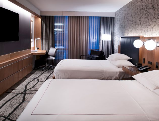 A modern hotel room with two beds, a wall-mounted TV, a desk with a chair, bedside tables, and contemporary decor, featuring a patterned wall.