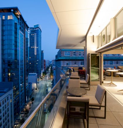 A modern cityscape at dusk, showcasing a high-rise balcony with seating and tables overlooking the street below and adjacent buildings.