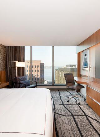 A modern hotel room with a large bed, wall-mounted TV, desk, chair, and a large window offering a city view.