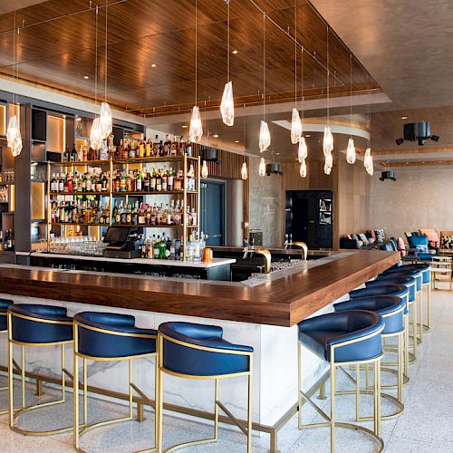 A modern bar with a fully stocked counter, blue and gold bar stools, hanging lights, and a seating area in the background.