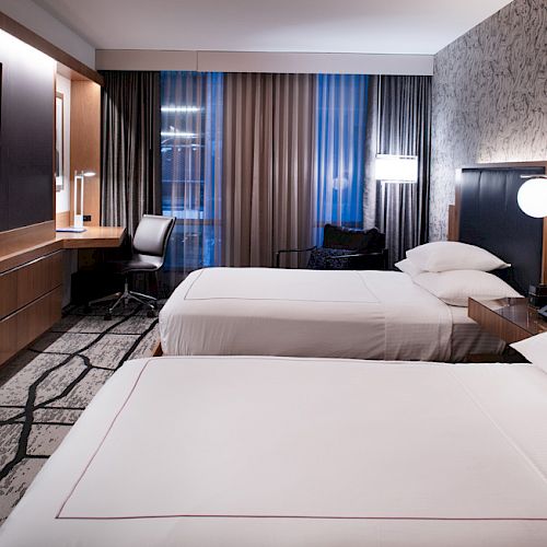 A modern hotel room with two double beds, a flat-screen TV, a work desk, and contemporary furnishings, illuminated by soft, ambient lighting.