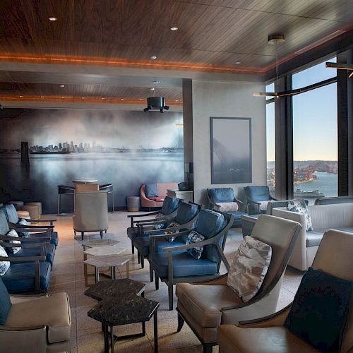 This image shows a lounge room with modern seating, large windows offering a scenic view, and wall art featuring a cityscape.