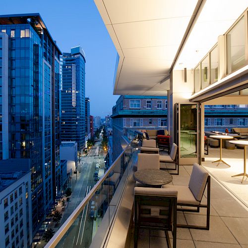 The image shows a modern rooftop terrace with seating and tables, overlooking a cityscape with tall buildings and streets at dusk.