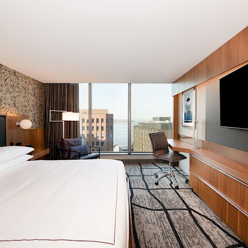 A modern hotel room with a large bed, desk, chair, and TV. A window offers a view of nearby buildings and a water body in the background.